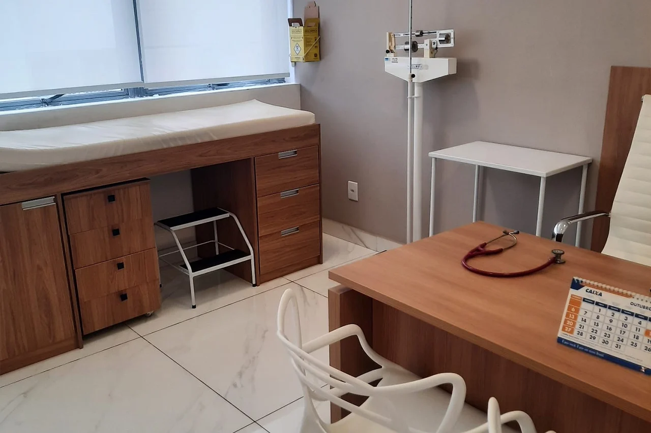 Consultório médico da reumatologista Dra. Larissa Karkow em Uberaba. O ambiente climatizado conta com mesa de atendimento de madeira, estetoscópio, cadeiras brancas, balança médica e maca para exames físicos presenciais.