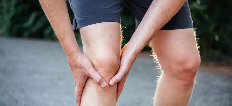 Homem com as mãos no joelho sentindo dor articular intensa provocada por sintomas de bursite ou tendinite.
